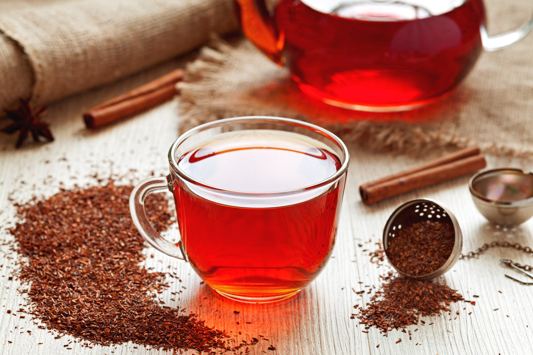 Rooibos Tee aufgebrüht in einer Glastasse (rote Färbung), drumherum loser Rooibos Tee